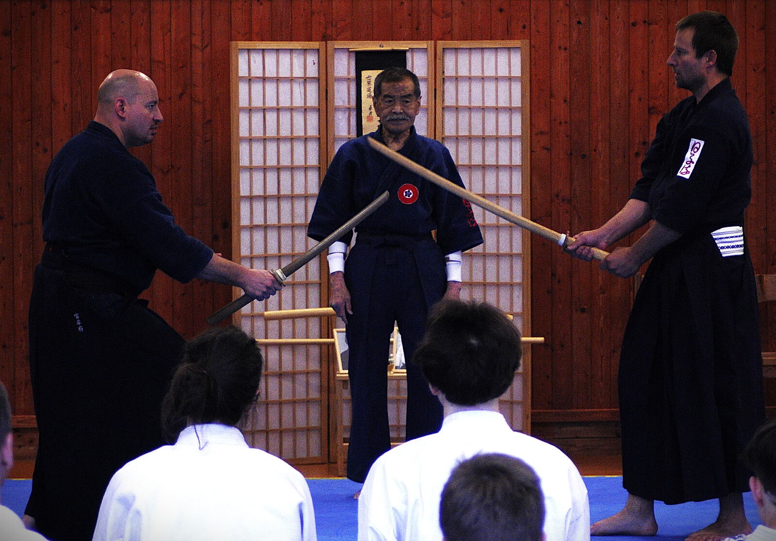 Seminář Hiko-ryu Taijutsu v dojo — dva cvičenci s dřevěnými meči, v popředí sedící žáci