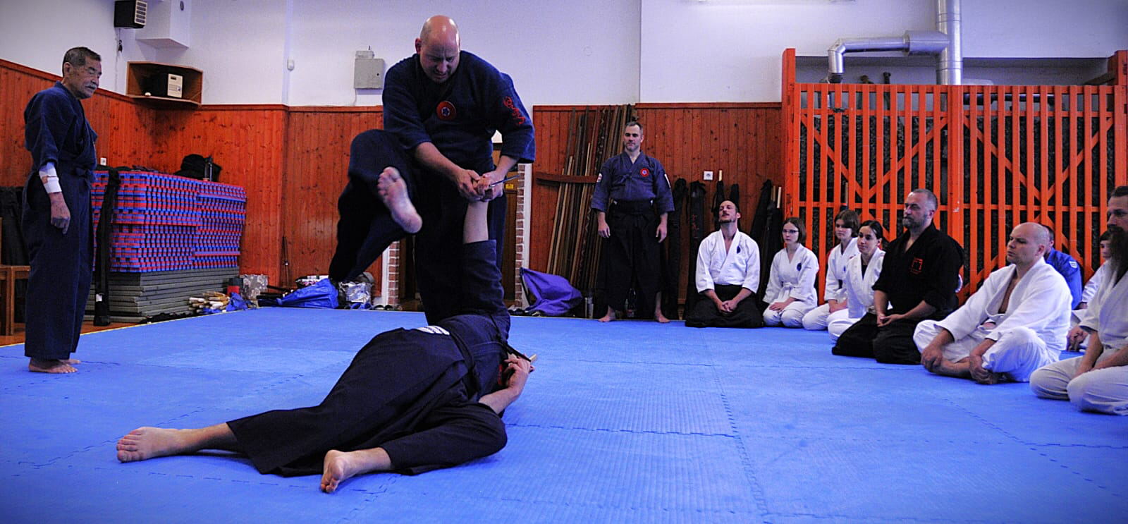 Seminář Hiko-ryu Taijutsu v dojo — kontrola ležícího útočníka s nožem, žáci sedí podél stěny